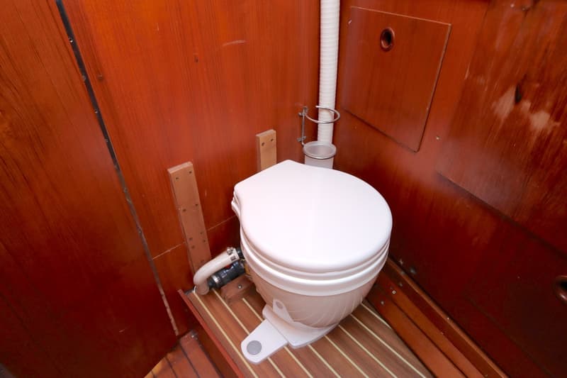 Een wit scheepstoilet met een pompmechanisme staat op een houten vloer met strepen, omringd door houten wanden.