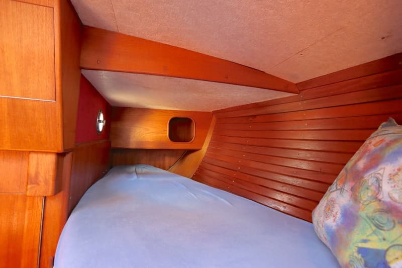 Een kajuit met een bed met lichtblauw hoeslaken, een kleurrijk kussen, houten wanden en een patrijspoort. — Koopmans 36