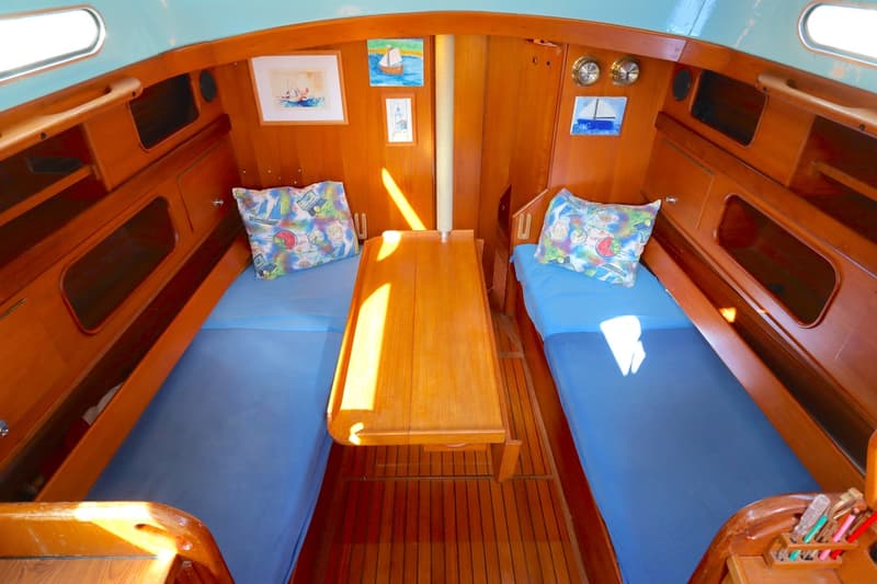 De foto toont het interieur van een boot met twee bedden met blauwe lakens en kussens, een houten tafel en houten wanden.