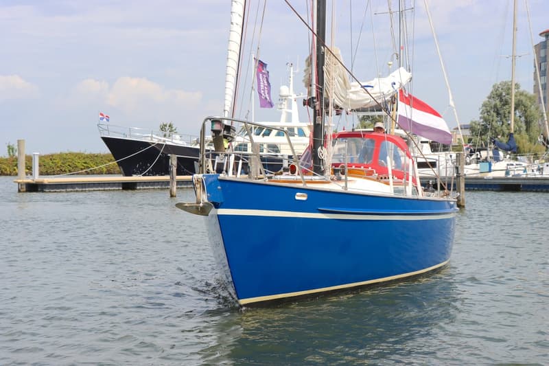 Een blauwe zeilboot met een rode buiskap en een Nederlandse vlag vaart op het water, met andere boten op de achtergrond.