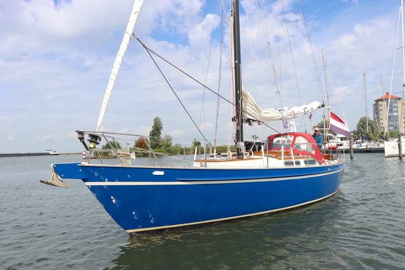 Een blauwe zeilboot met een rode buiskap en een Nederlandse vlag, liggend in het water onder een bewolkte hemel.