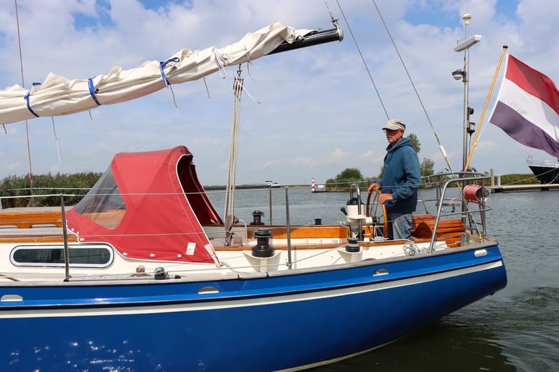 Een man staat aan het roer van een blauw-witte zeilboot met een rode buiskap en een opgerold zeil. — Koopmans 36