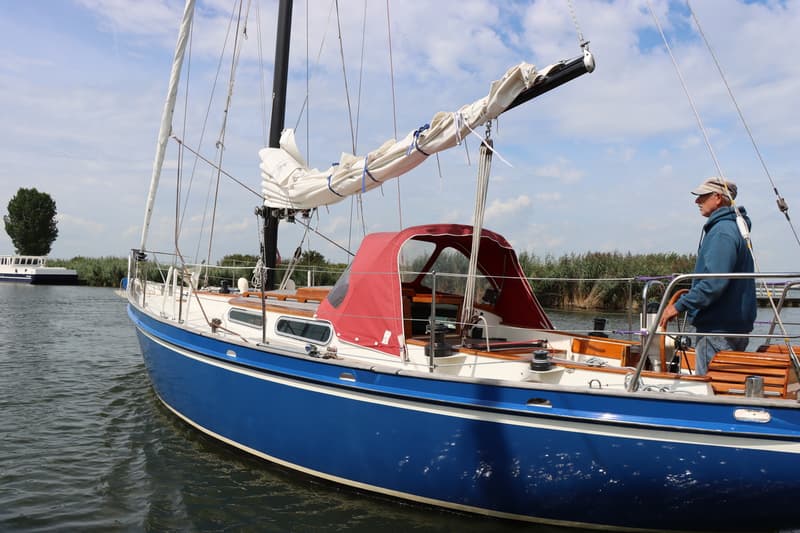 Een blauwe zeilboot met een rode buiskap en een opgerold zeil ligt in het water, met een man aan het roer. — Koopmans 36