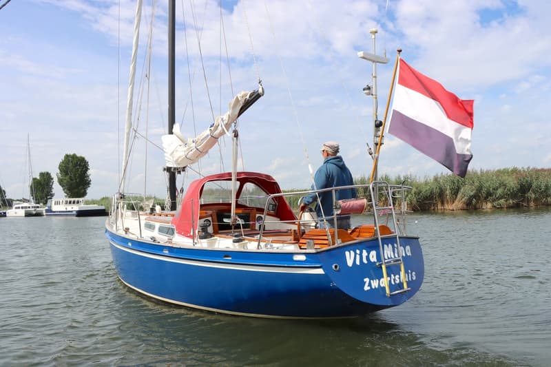 Een blauwe zeilboot met een rode buiskap en een Nederlandse vlag vaart op het water, met een persoon aan het roer.