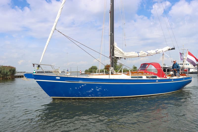Een blauwe zeilboot met een rode buiskap en een opgerold grootzeil vaart op het water met een persoon aan het roer.