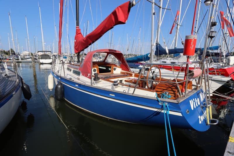 Een blauwe zeilboot met rode zeilhuik en buiskap ligt afgemeerd in een haven vol met andere zeilboten. — Koopmans 36