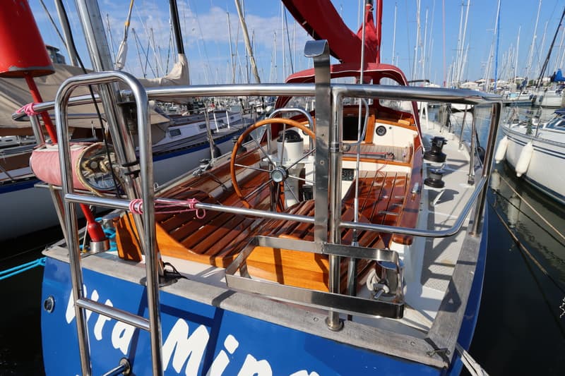 Een blauwe zeilboot met een houten stuurwiel en een rood zeil, omringd door andere boten in een haven. — Koopmans 36