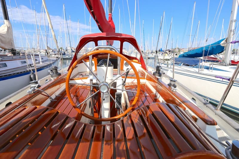 Een houten stuurwiel en een kompas op een zeilboot met een rode kap, omringd door andere zeilboten in een haven.