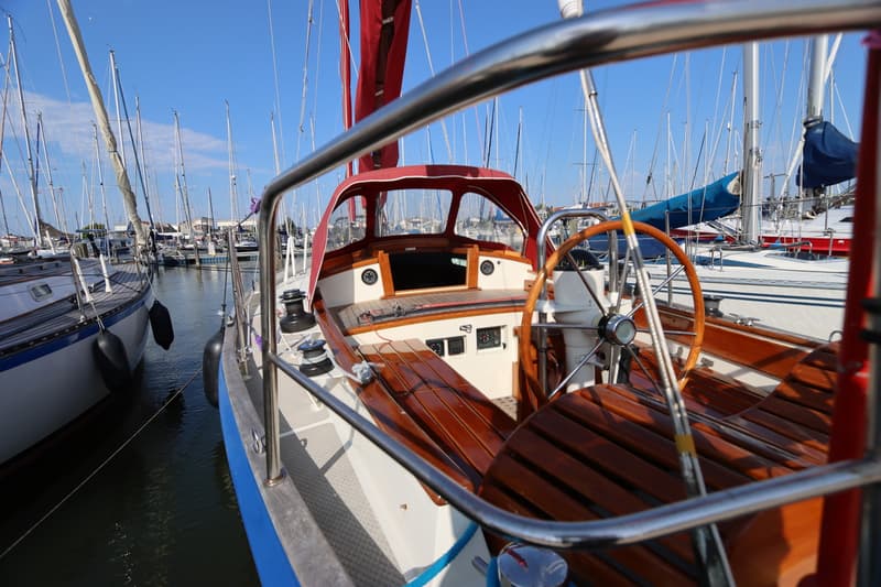 Een houten stuurwiel en een rood zeil zijn zichtbaar op een boot in een haven vol zeilboten. — Koopmans 36