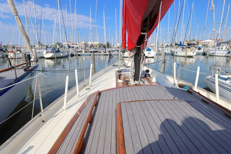 Een bovenaanzicht van het dek van een zeilboot met een rood zeil, liggend in een jachthaven vol met andere boten.