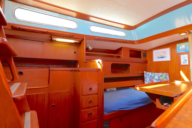 De foto toont het interieur van een boot met houten betimmering, een bed met blauw laken en diverse opbergvakken.