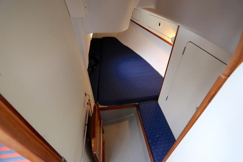 Een interieur van een boot met een blauw gestippeld matras en kussens, omringd door witte wanden en houten accenten.