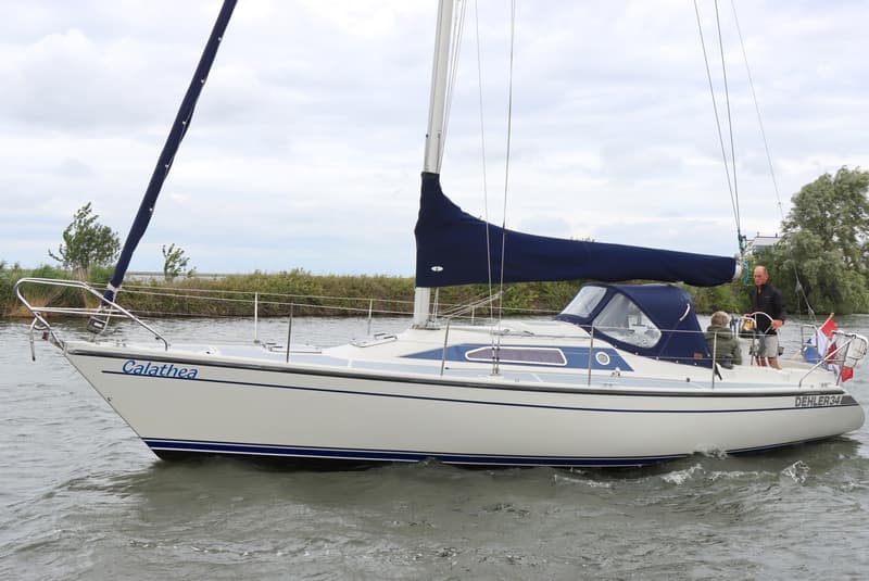 Een witte zeilboot met blauwe accenten en de naam "Calathea" vaart op water, met twee personen aan boord.