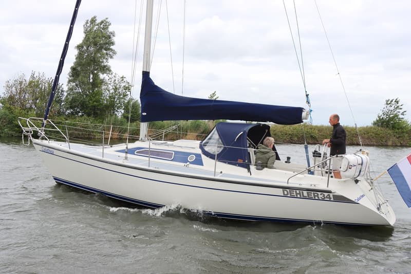Een witte zeilboot met blauwe accenten en de naam "Calathea" en "DEHLER 34" vaart op water met twee personen aan boord.