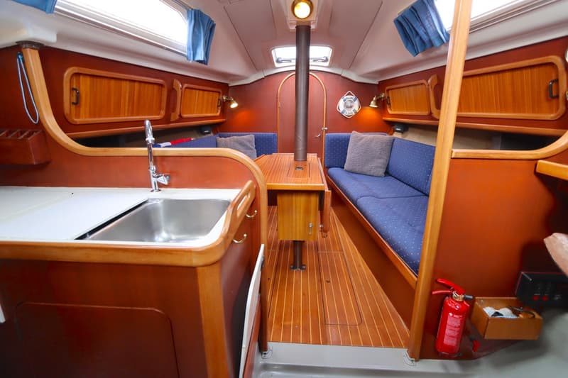 De foto toont het interieur van een boot met een kombuis aan de linkerkant en twee banken met een tafel in het midden.