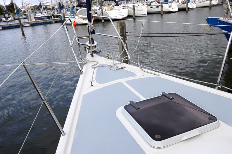 De foto toont het voordek van een zeilboot met een luik en reling, liggend in een haven met andere boten.