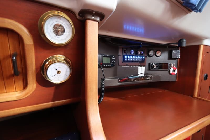 Een interieur van een boot met houten panelen, een barometer, een klok, een marifoon, een schakelpaneel en een radio.