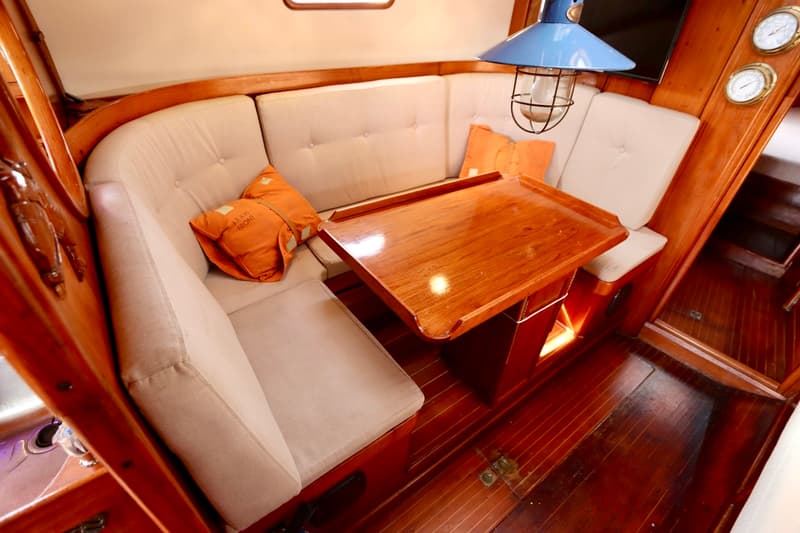 De foto toont een kajuitinterieur met een U-vormige bank met lichte kussens, een houten tafel en een blauwe lamp.