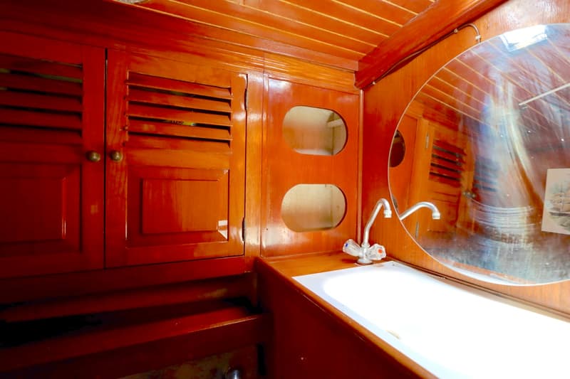 Een houten interieur met een wasbak, twee kranen, een ronde spiegel en kasten met jaloezieën en open vakken.
