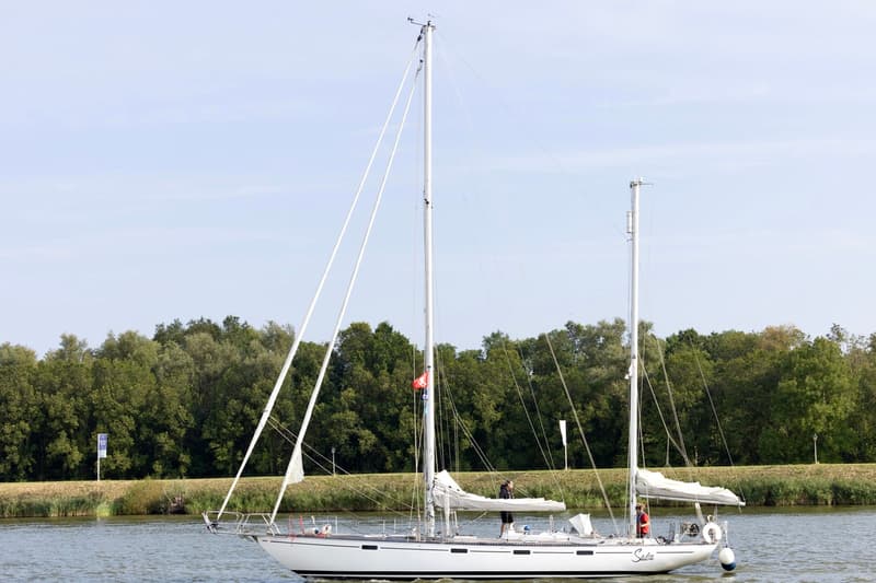 Een witte zeilboot met twee masten vaart op een waterweg, met een bosrijke oever op de achtergrond. — RJ 140