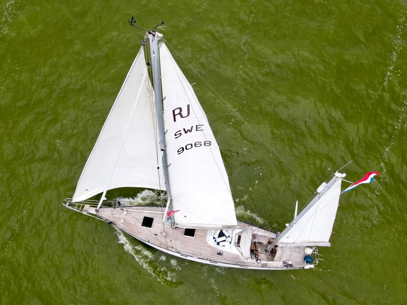 Een zeilboot met witte zeilen en een houten dek vaart op groen water, gezien van bovenaf. — RJ 140
