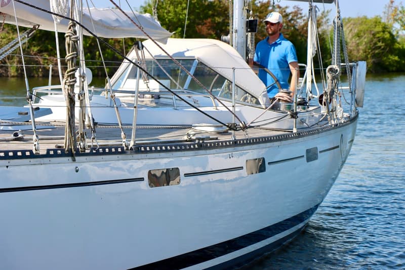 Een man in een blauw poloshirt en witte pet staat aan het roer van een witte zeilboot met een houten dek. — RJ 140