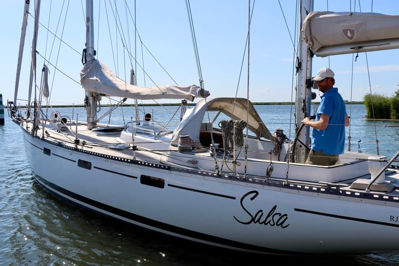 Een witte zeilboot met de naam "Salsa" op de romp, met een man in een blauw shirt aan het roer. — RJ 140