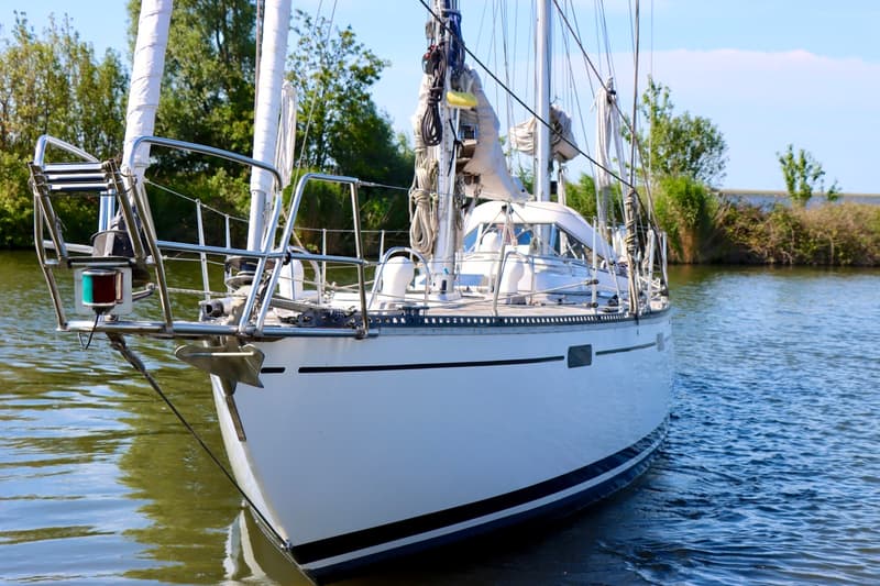 Een witte zeilboot met een zwarte en blauwe streep op de romp ligt in het water, met bomen op de achtergrond. — RJ 140