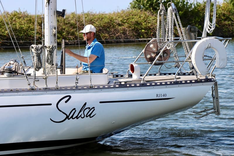 Een witte zeilboot genaamd "Salsa" met een man aan boord vaart op water met begroeiing op de achtergrond. — RJ 140