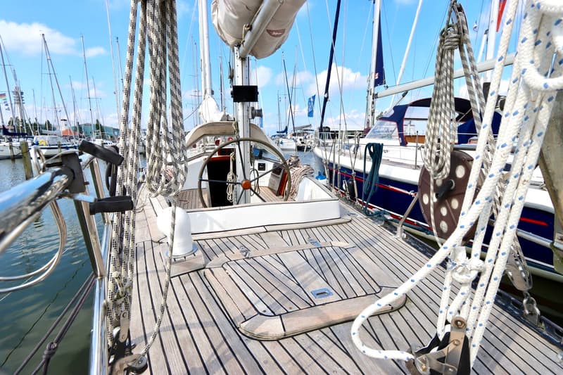 Een houten dek met een stuurwiel, touwen en een lier, omringd door andere boten in een haven.