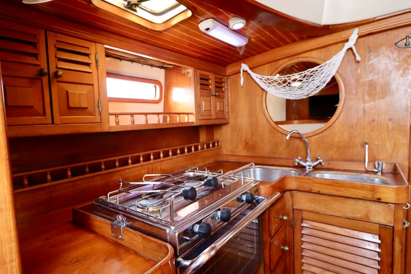 De foto toont een houten scheepskeuken met een fornuis, dubbele spoelbak, kasten en een rond raam met een visnet.