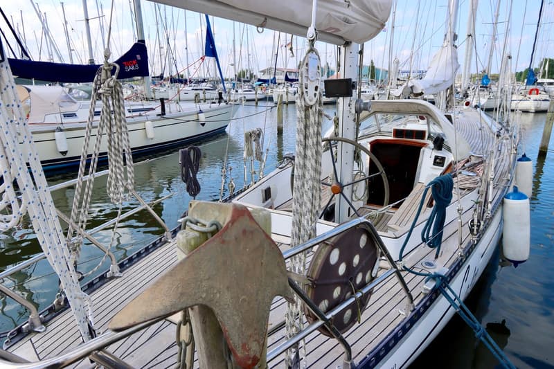 Een witte zeilboot met een teakhouten dek ligt afgemeerd in een haven vol met andere zeilboten. — RJ 140