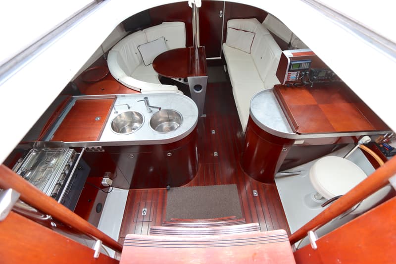 De foto toont het interieur van een boot met een kombuis, zithoek en navigatiehoek, alles afgewerkt met donker hout en…