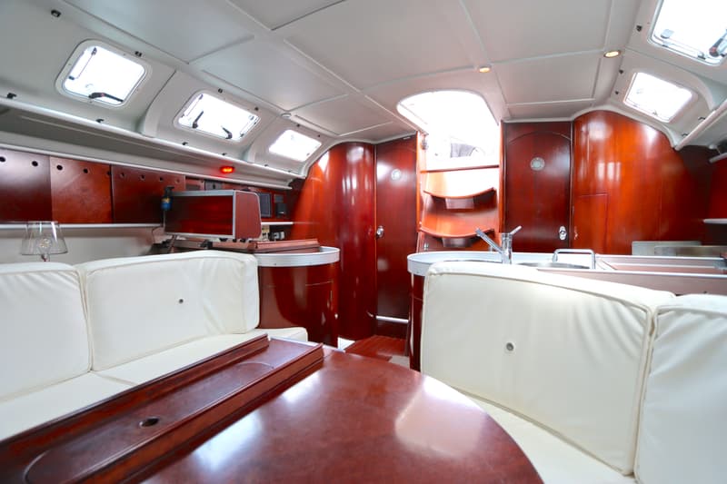 De foto toont het interieur van een boot met houten wanden, witte banken, een tafel en een kombuis met spoelbakken.