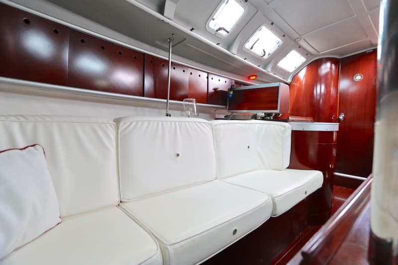 De foto toont een interieur van een boot met witte banken, houten kasten en een aantal ramen in het plafond.