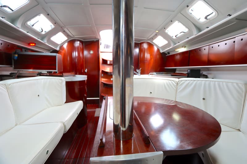 De foto toont het interieur van een boot met een houten vloer, witte banken, een ronde houten tafel en een zilverkleurige…