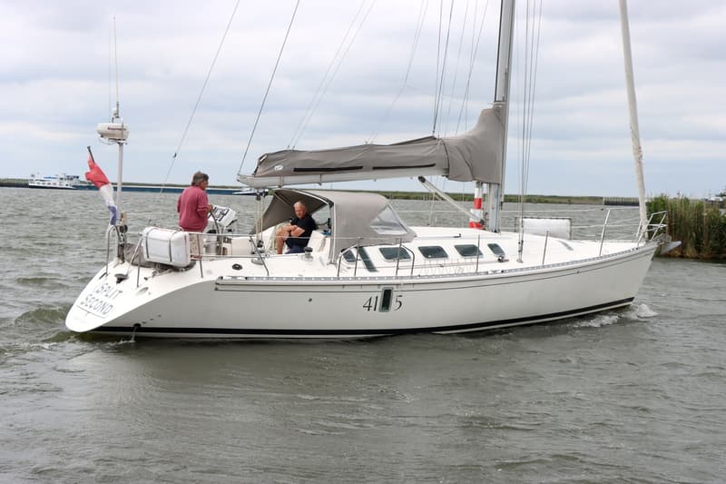 Een witte zeilboot met twee mannen aan boord vaart op water, met een Nederlandse vlag aan de achtersteven.