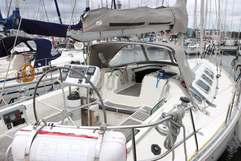 Een witte zeilboot met een stuurwiel, navigatie-instrumenten en een grijze buiskap, liggend in een jachthaven.