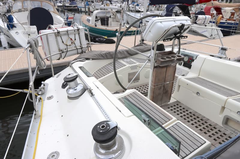 Een witte zeilboot ligt aangemeerd in een haven, met een stuurwiel, lieren en een reddingsvlot zichtbaar.