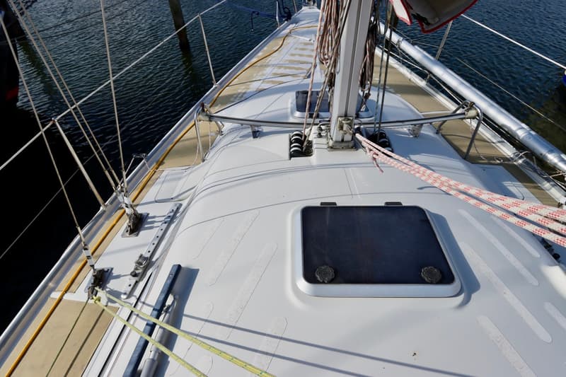 Een bovenaanzicht van het dek van een zeilboot met een mast, touwen, luiken en relingen, liggend aan het water.