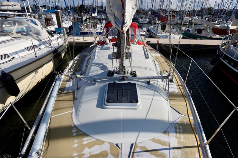 Een bovenaanzicht van het dek van een zeilboot met een opgerold zeil en een luik, omringd door andere boten in een haven.
