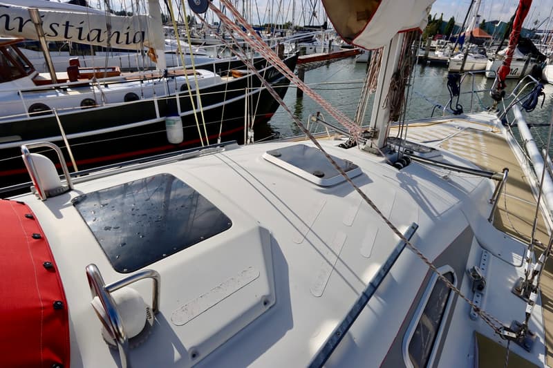Een bovenaanzicht van het dek van een zeilboot met twee luiken, touwen en een rode huik, liggend in een haven.