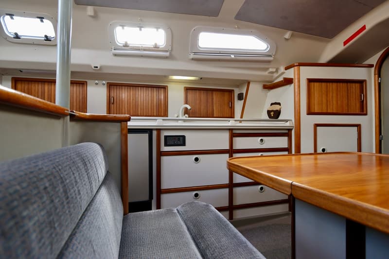 De foto toont het interieur van een boot met een zithoek, een kombuis met wasbak en een houten tafel. — Dehler 36 CWS