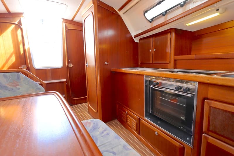De foto toont een houten bootinterieur met een kombuis inclusief oven/fornuis, een tafel met zitbanken en een raam.