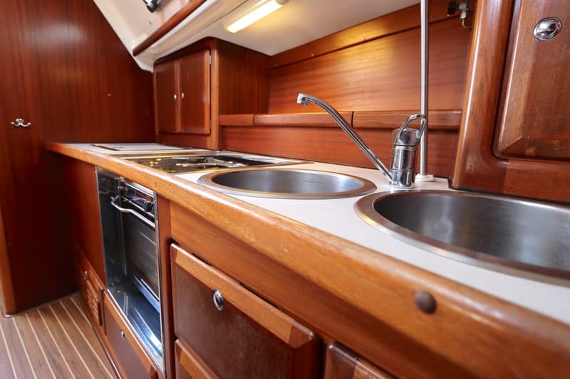 De foto toont een houten scheepsinterieur met een witte aanrecht, twee roestvrijstalen spoelbakken, een kraan en een oven.