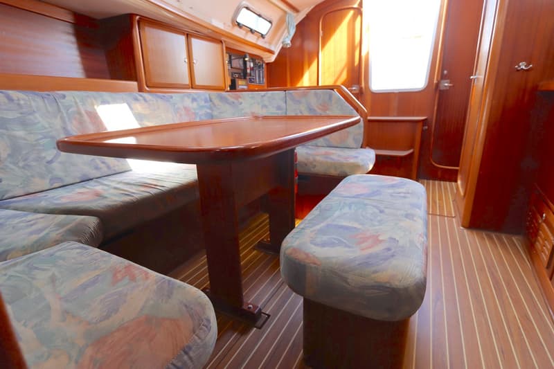 Een houten kajuit met een tafel en banken met lichtblauwe, roze en groene kussens, en een houten vloer. — Bavaria 35 Holiday