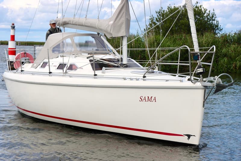 Een witte zeilboot met de naam "SAMA" en een rode streep op de romp vaart op het water, met een persoon aan boord. — Etap 30i