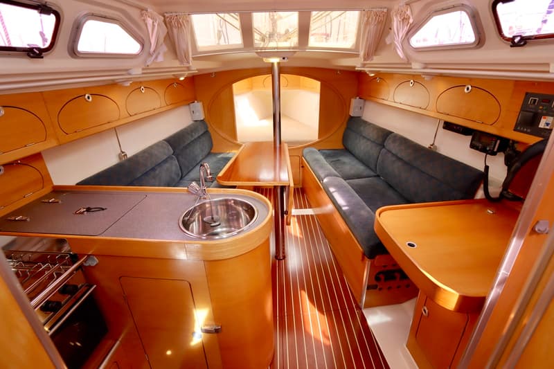 Het interieur van de boot toont een kombuis met gootsteen en fornuis, een zithoek met blauwe kussens en een houten tafel.