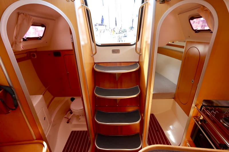 De foto toont het interieur van een boot met een centrale trap, een toiletruimte links en een slaapcabine met kombuis rechts.