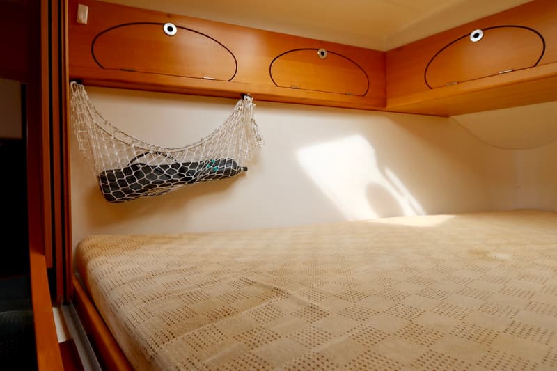 Een kajuit met een bed met lichtbruine sprei, daarboven houten kasten en een wit net met een zwart voorwerp. — Etap 30i